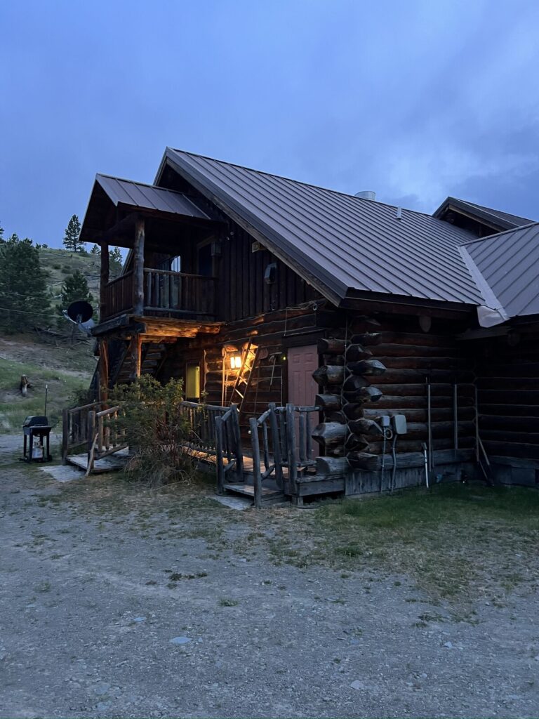 Two-story log lodge wing with an upper balcony and warm porch light at dusk