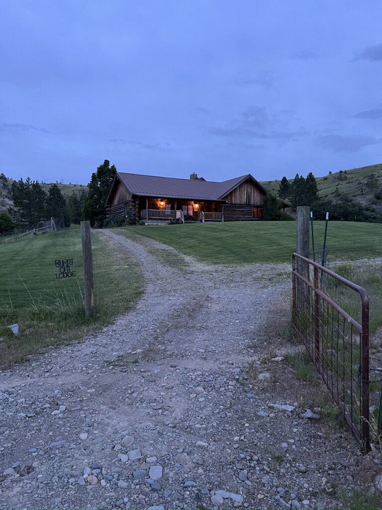 Gravel drive and ranch gate leading up to the lit Spring Creek Lodge at dusk