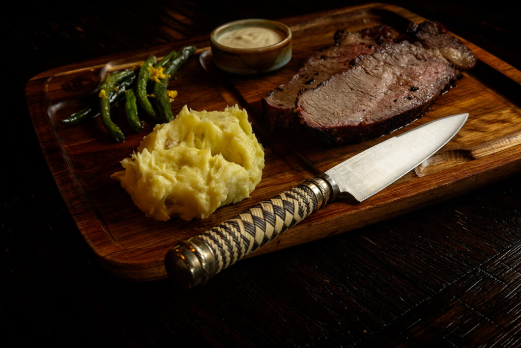 Grilled steak, mashed potatoes, blistered green beans, and a creamy sauce plated on a wood board